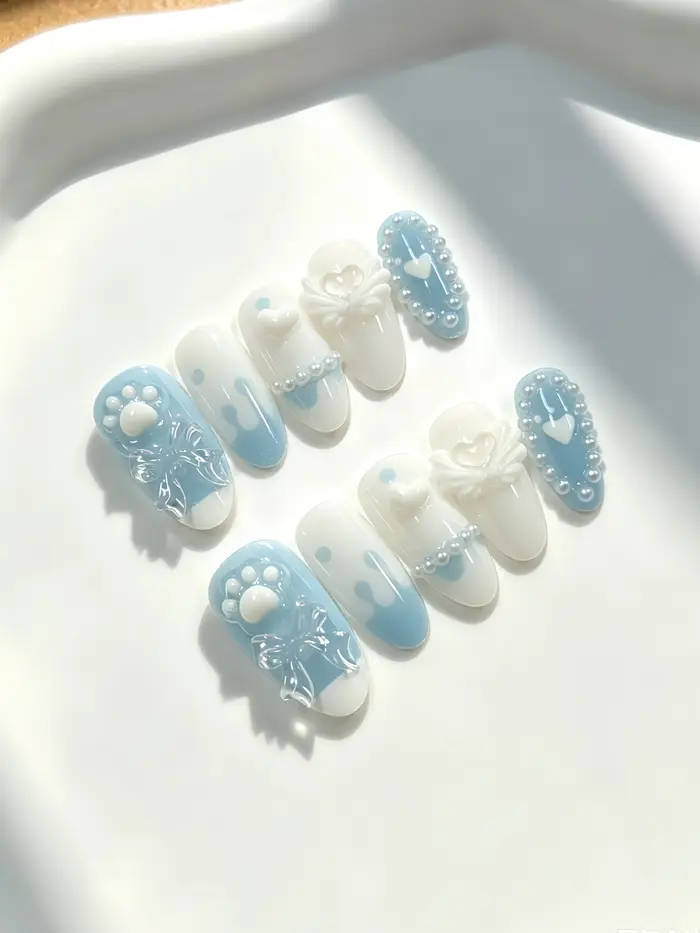 10 ongles de longueur moyenne en forme d'amande, aux couleurs principales blanc laiteux et bleu clair, décorés de nœuds transparents blanc laiteux en forme de patte de chat, d'ailes en forme de cœur,